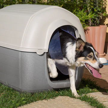 Dog exiting a gray pet house on grass with a garden in the background